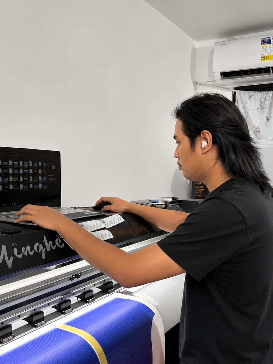 Long-haired operator at a laptop beside a wide-format sublimation printer